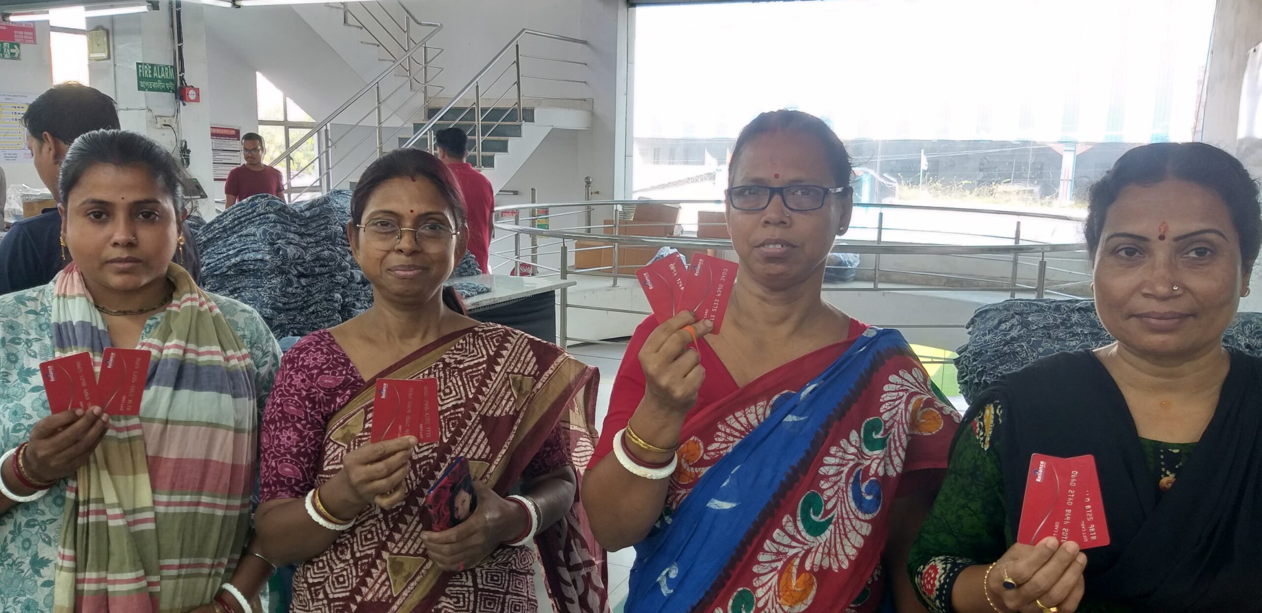 Four female garment workers wearing colorful saris stand indoors, each holding red Fair Trade premium cards.