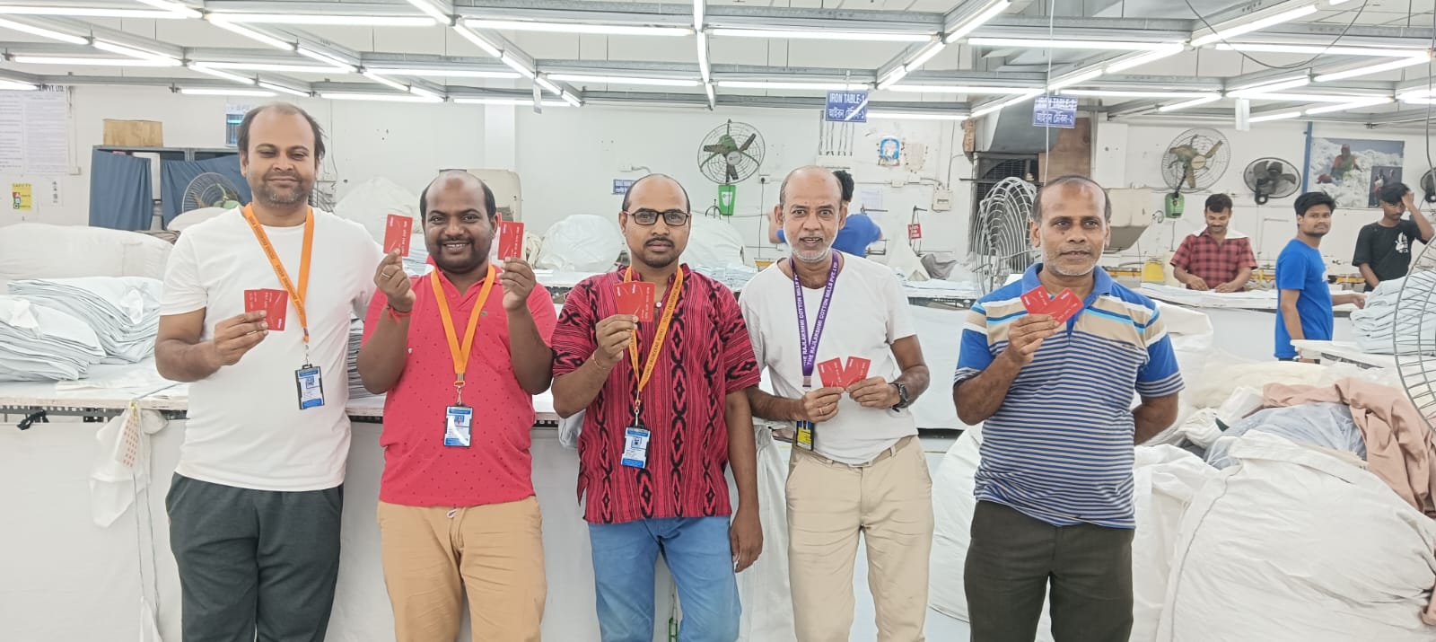 Six male garment workers standing inside a factory, smiling and holding red cards, with folded clothes and fans in the background.