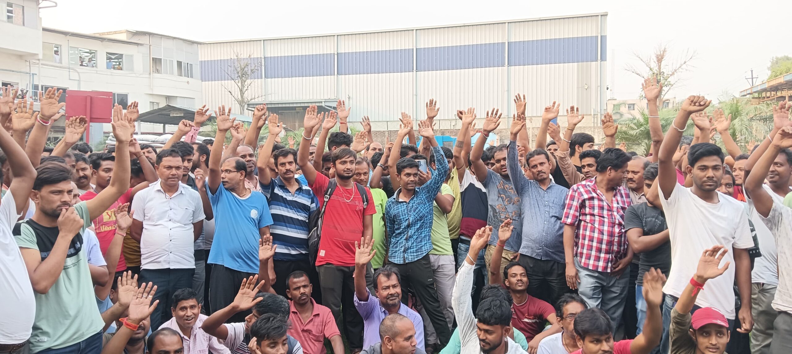 A large group of male workers gathered outdoors, raising their hands in solidarity during a meeting outside a factory building.